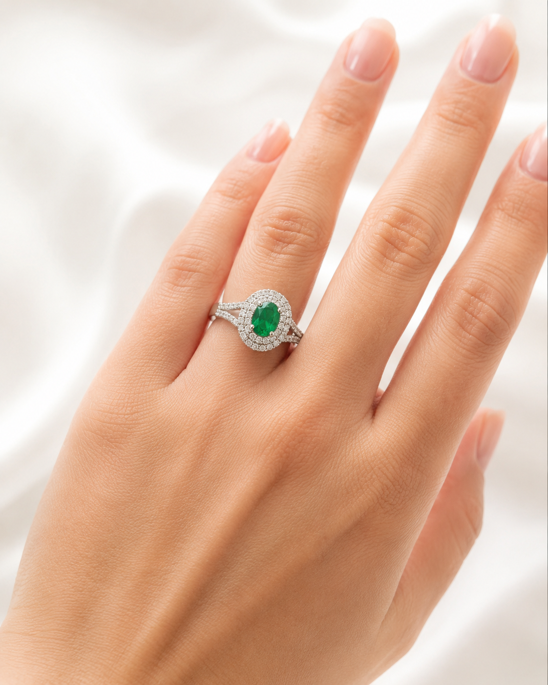 A close-up of a hand wearing a silver-toned halo ring with a central oval green emerald gemstone and small surrounding clear stones, set against a soft white fabric background.