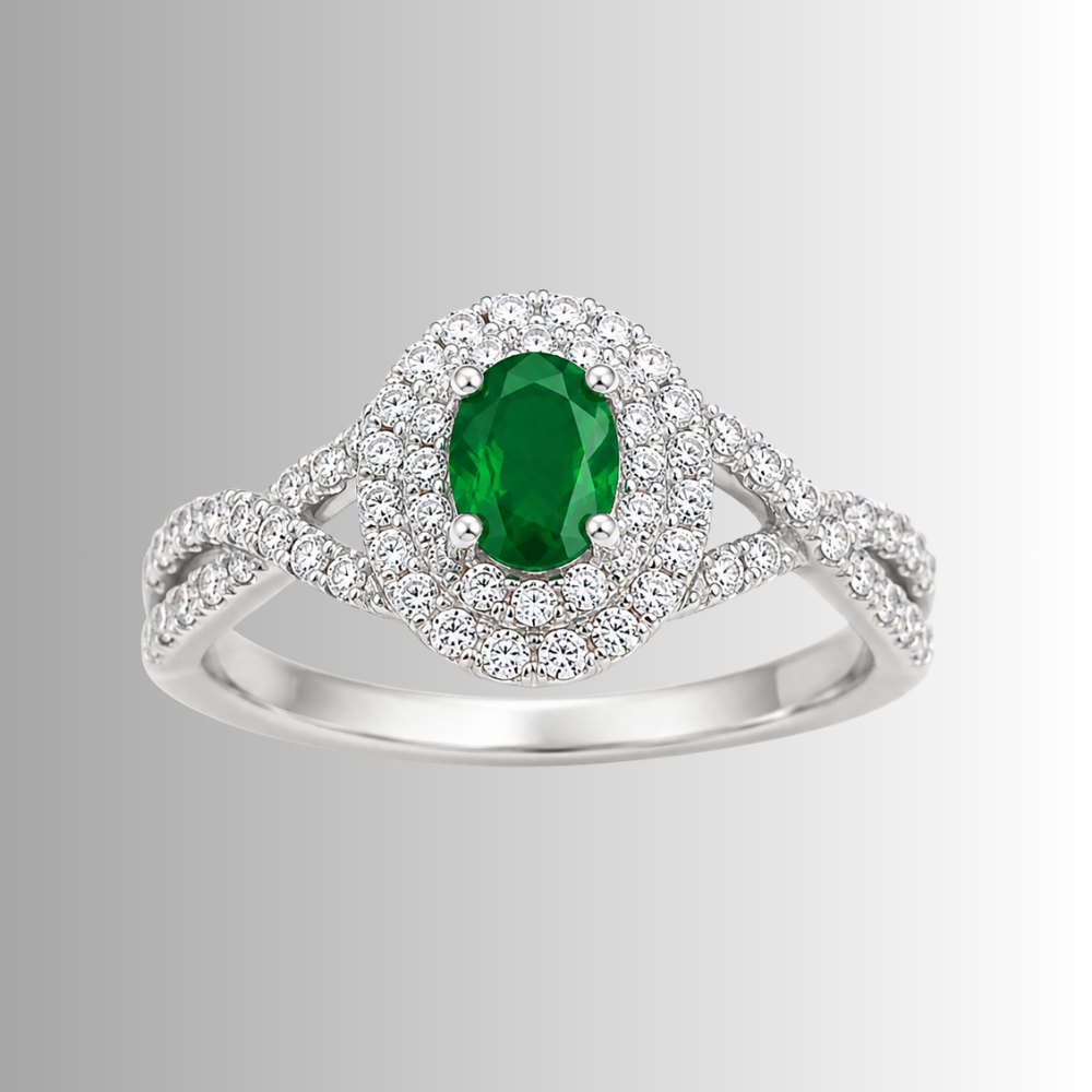 A silver-toned halo ring featuring an oval-cut green emerald center stone surrounded by a double halo of small clear stones, set on a split shank band, shown against a light gray gradient background.