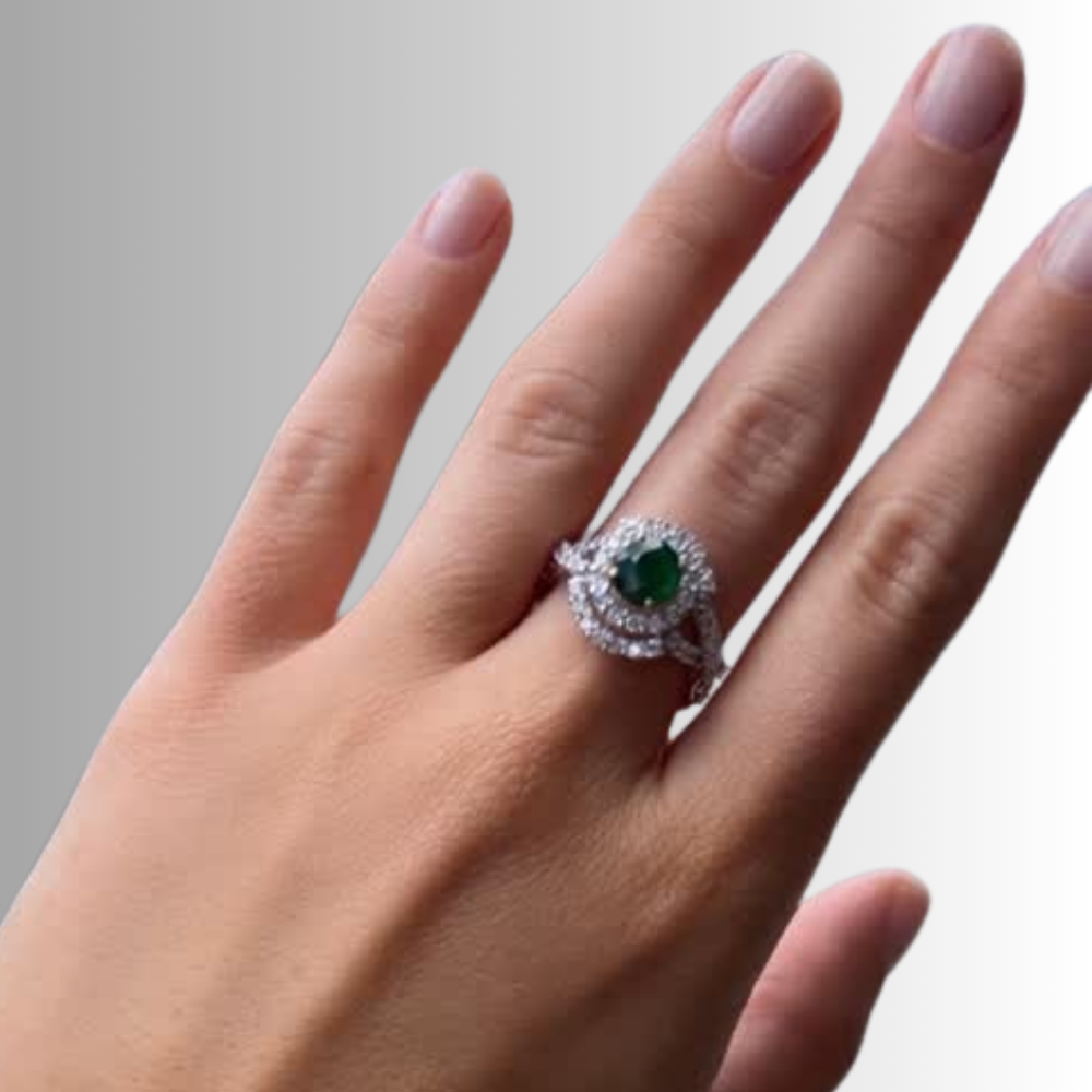 A close-up of a hand wearing a silver-toned halo ring with a round green emerald center stone and small clear accent stones.